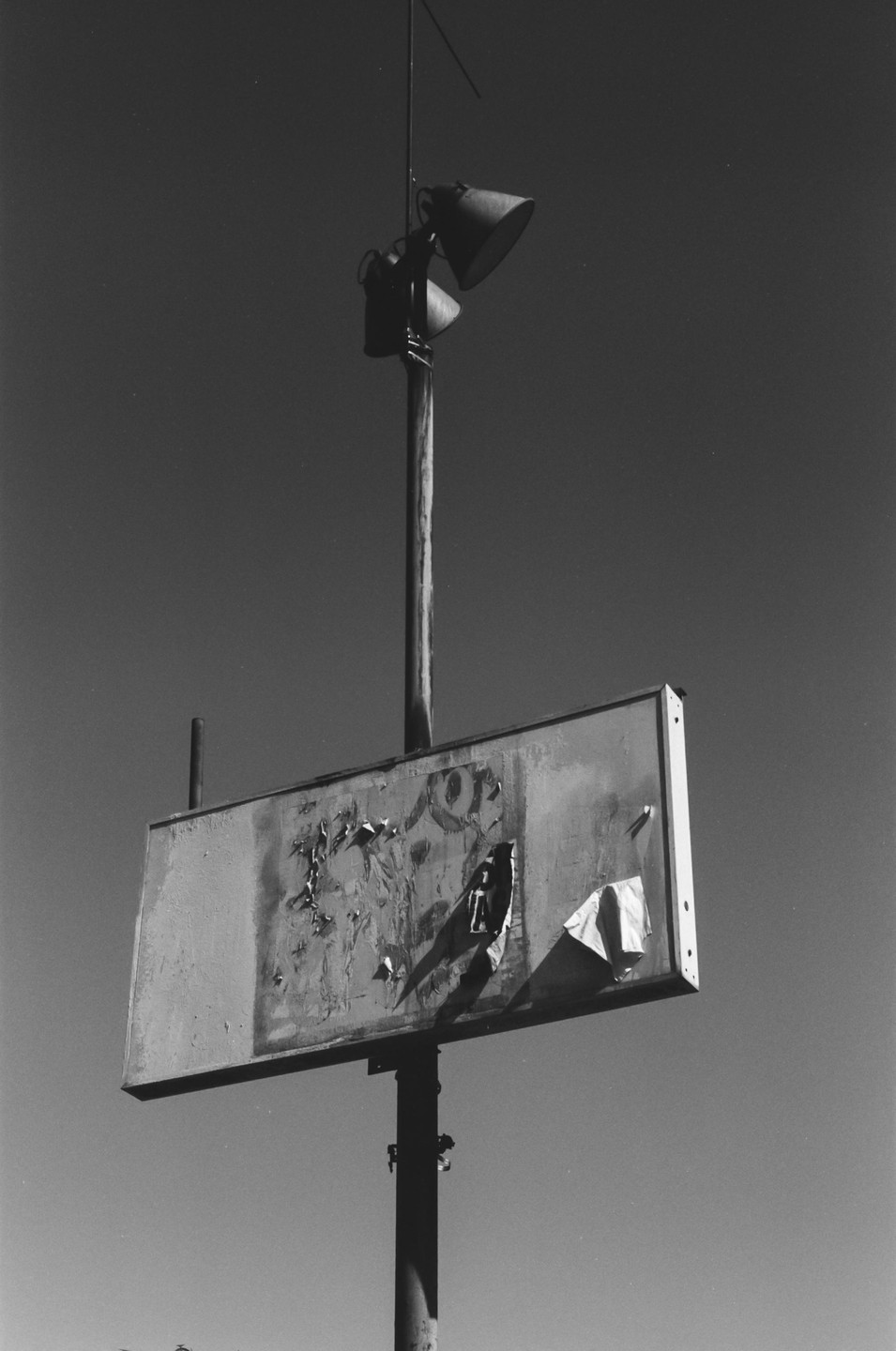 A weathered sign with peeling paper and two spotlights on a pole stands against a clear sky.