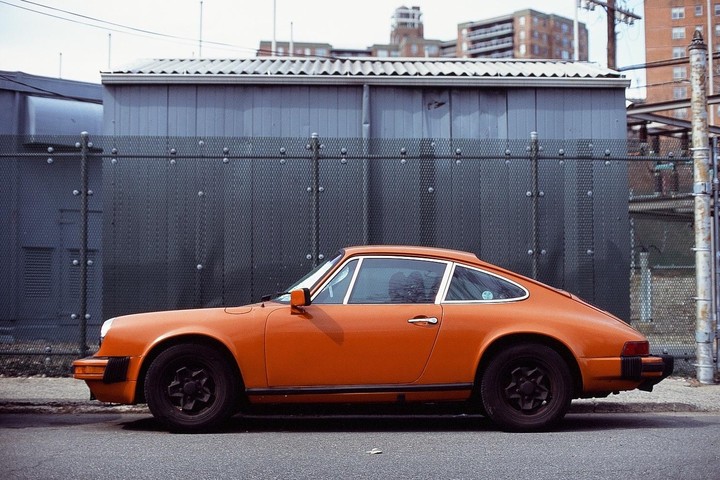 An orange sports car is parked on a street next to a metal fence with buildings in the background.