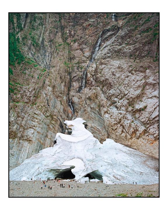 A large ice cave with people standing near its entrance is at the base of a rocky mountain with a waterfall.