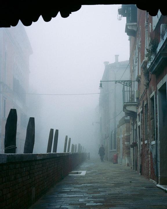 A person walks down a narrow, foggy street lined with buildings and a brick wall with wooden posts.