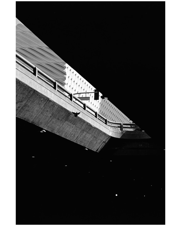 A bird flies under a concrete overpass next to a building with a textured facade.