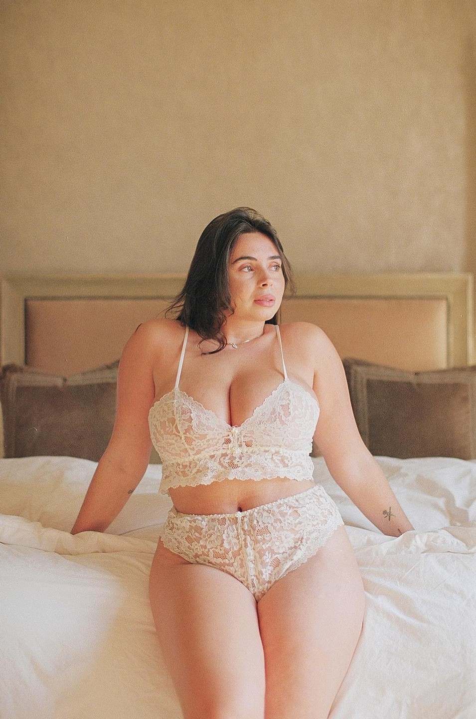 A woman wearing white lace lingerie sits on a bed and looks to the right.