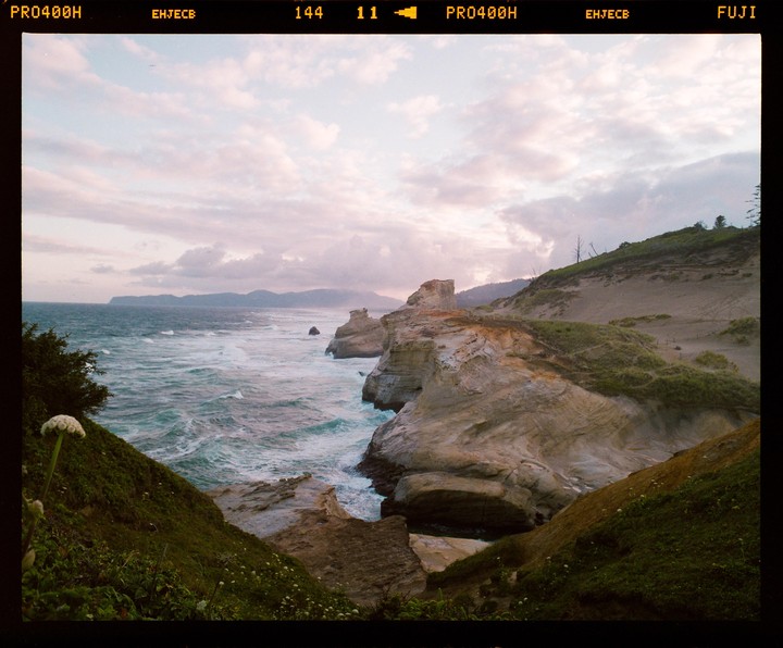 Waves crash against a rocky coastline under a pastel sky with a large sand dune on the right.