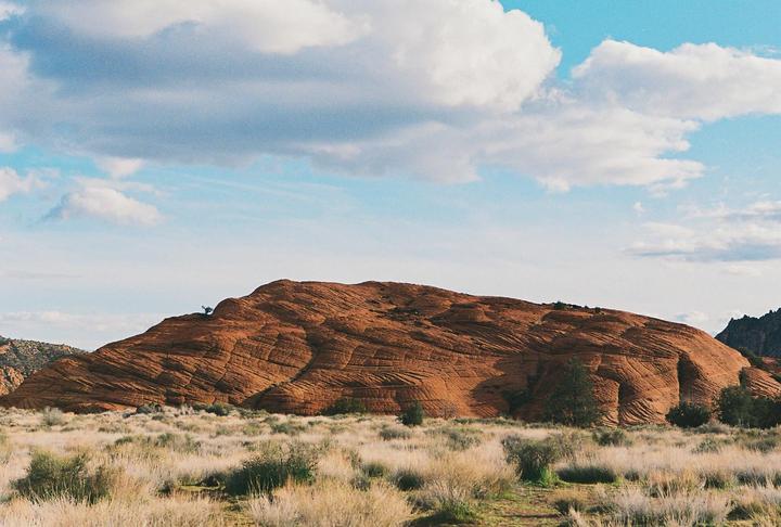 A large red rock formation with textured patterns rises from a desert landscape under a blue sky with white clouds.