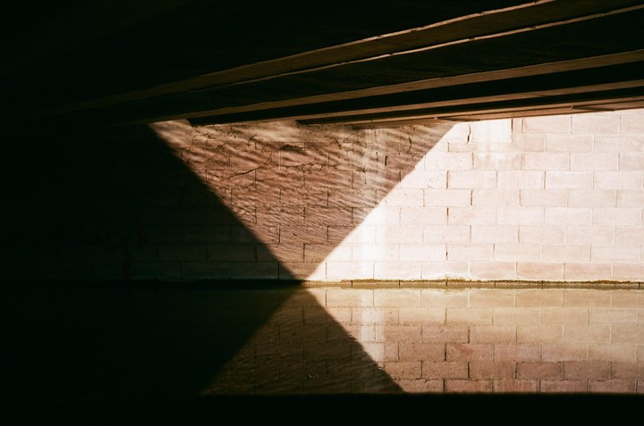 Sunlight creates a triangular pattern on a brick wall and its reflection in water under a dark overhang.