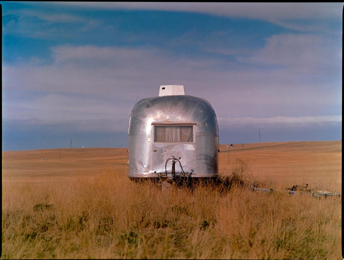 A silver travel trailer sits in a field of tall, dry grass under a partly cloudy sky.