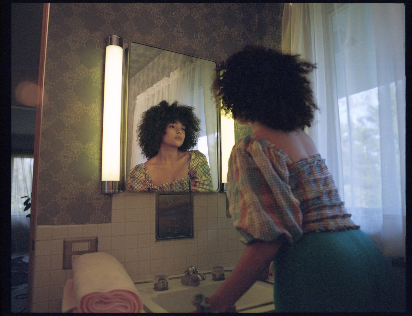 A woman with curly hair looks at her reflection in a mirror above a sink.