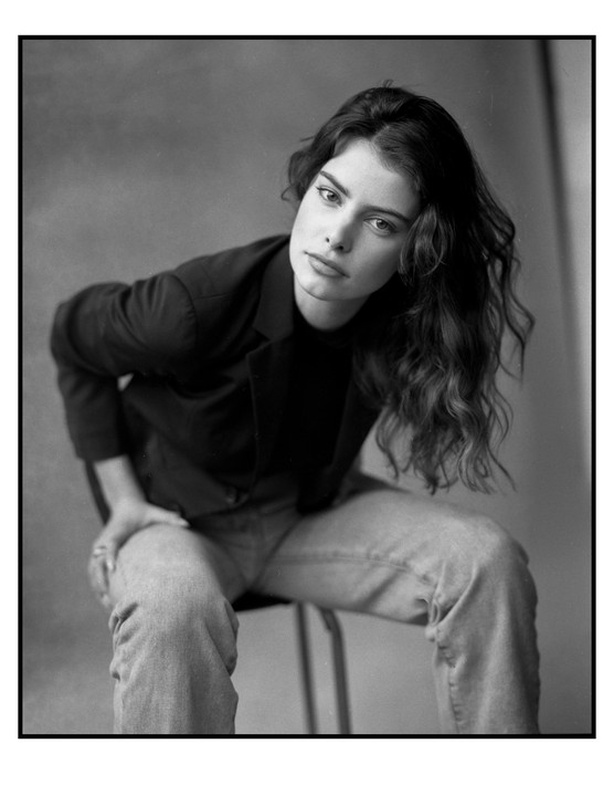 A woman with long, wavy hair sits on a stool, looking directly at the viewer.