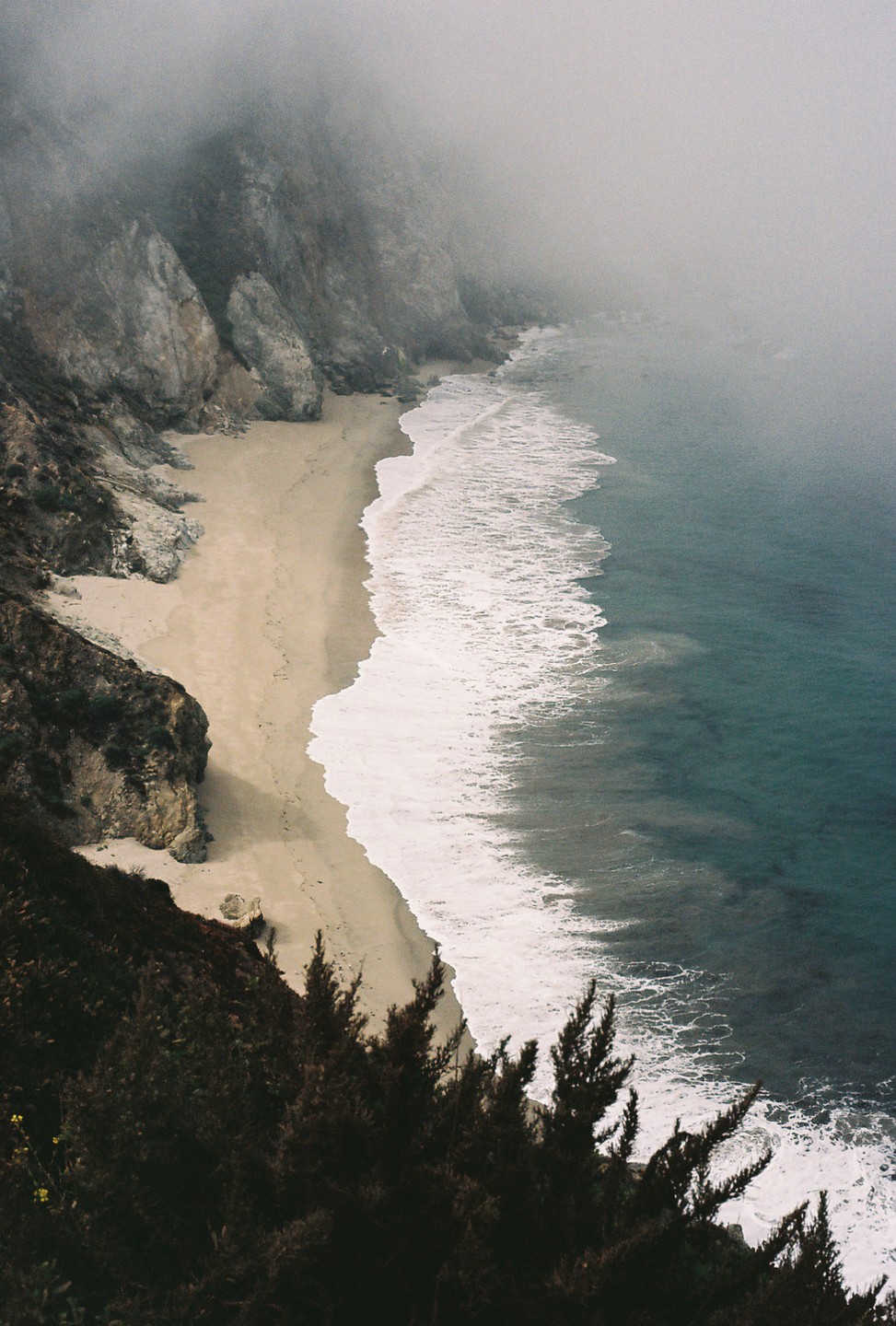 A sandy beach with waves crashing on the shore is seen from above, with a cliff face on the left and fog obscuring the top of the cliff and the horizon.
