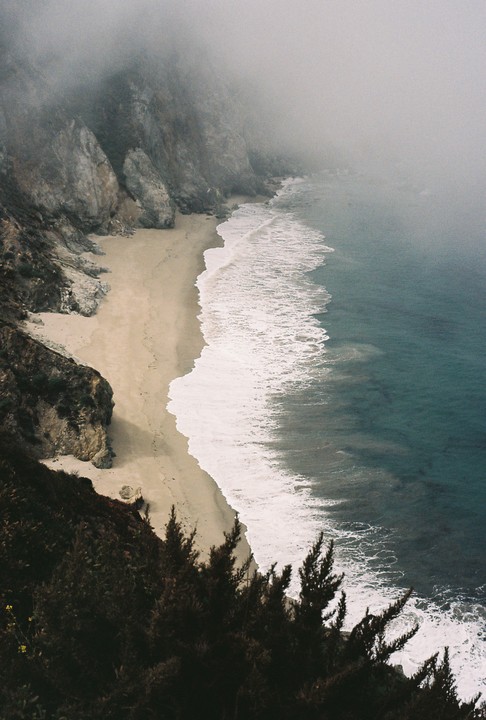 A sandy beach with waves crashing on the shore is seen from above, with a cliff face on the left and fog obscuring the top of the cliff and the horizon.