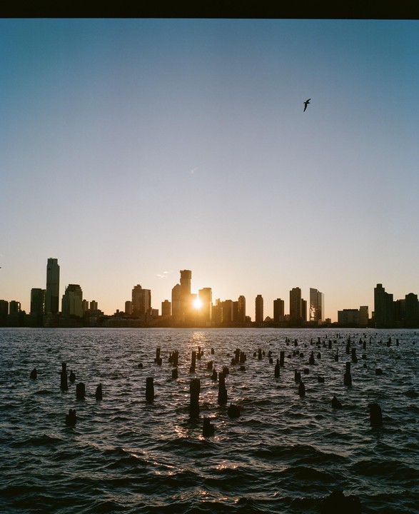 The sun sets behind a city skyline across a body of water with many wooden posts.