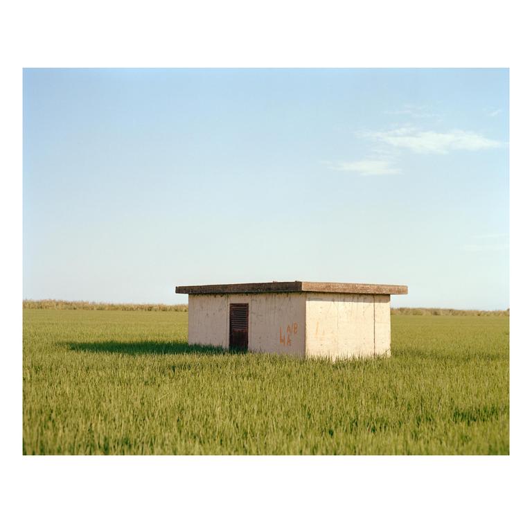 A small concrete building with a dark door stands in a field of green grass under a clear sky.