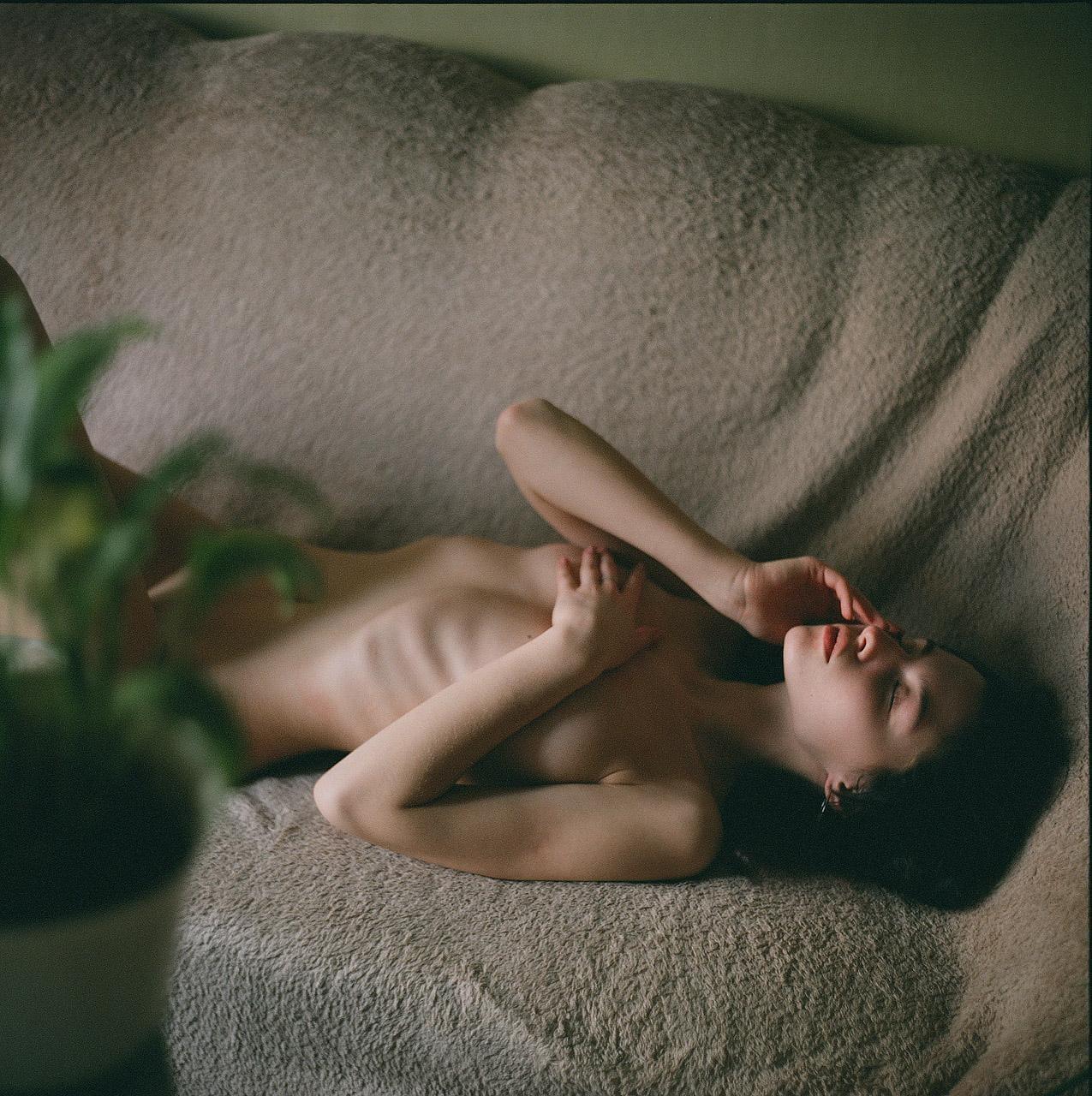 A nude woman lies on a textured surface with her eyes closed and hands near her face, with a plant in the foreground.