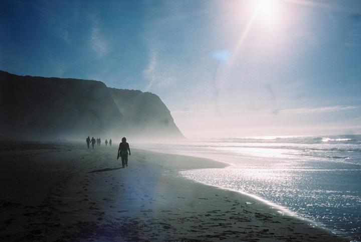 A group of people walk along a beach with cliffs in the background and bright sunlight reflecting on the water.