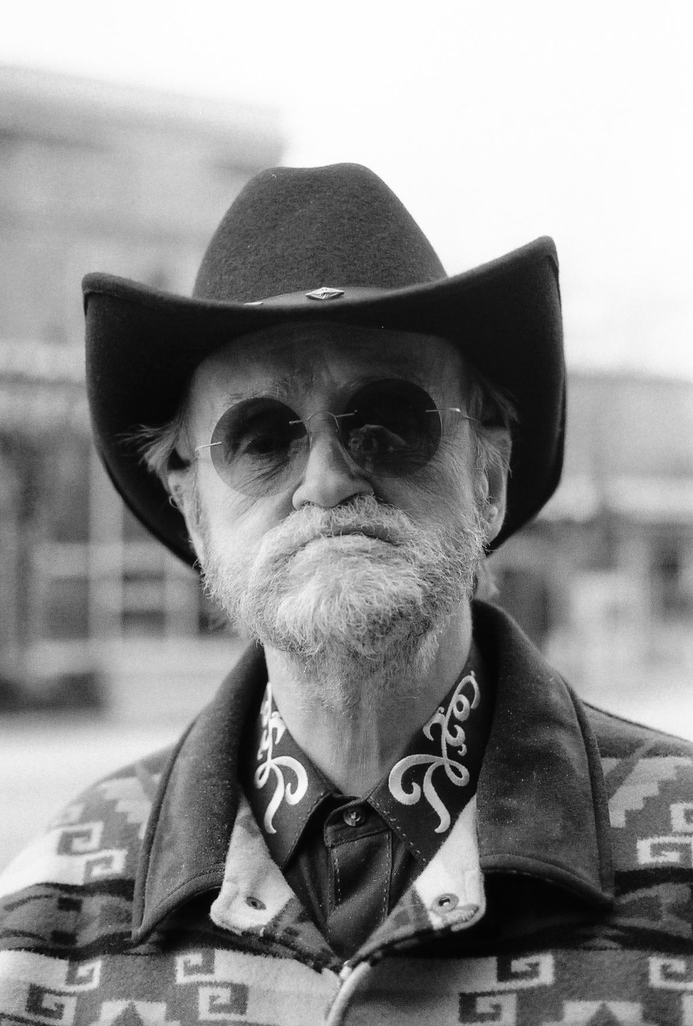 A man with a beard, sunglasses, cowboy hat, and patterned jacket looks directly at the viewer.
