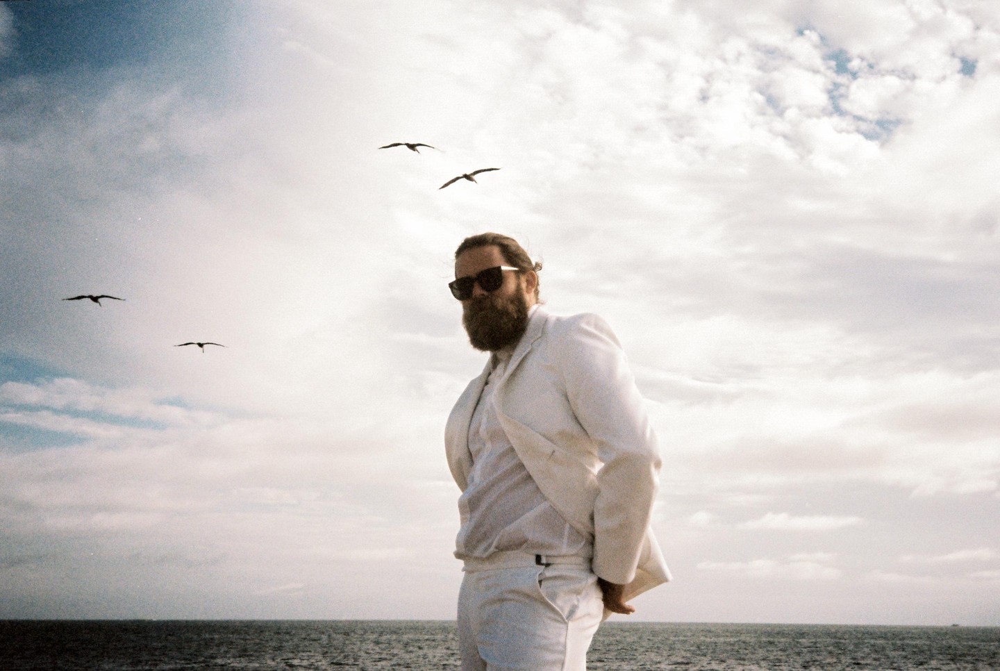 A man in a white suit and sunglasses stands by the ocean with birds flying overhead.