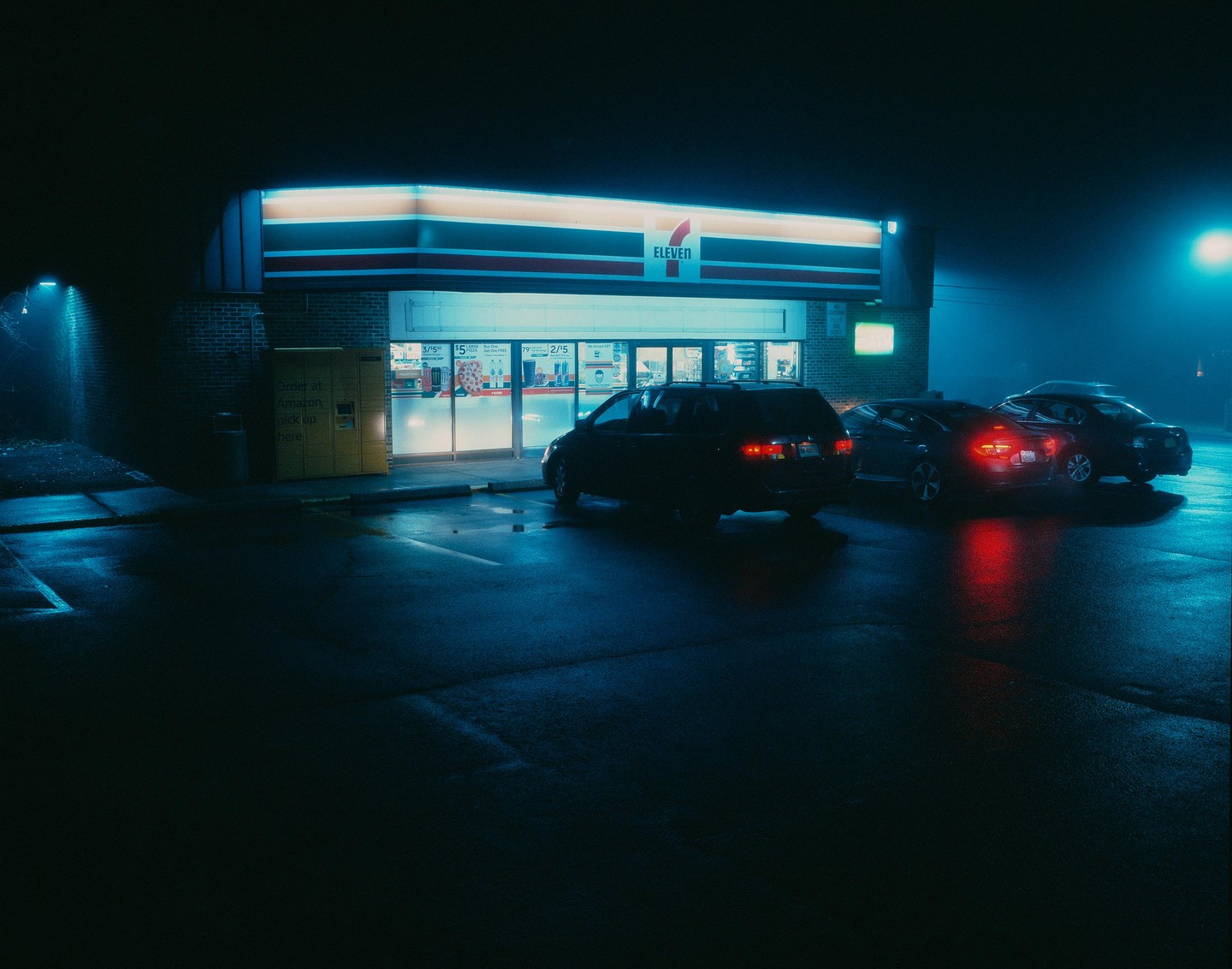 A 7-Eleven convenience store is illuminated at night with several cars parked in front.