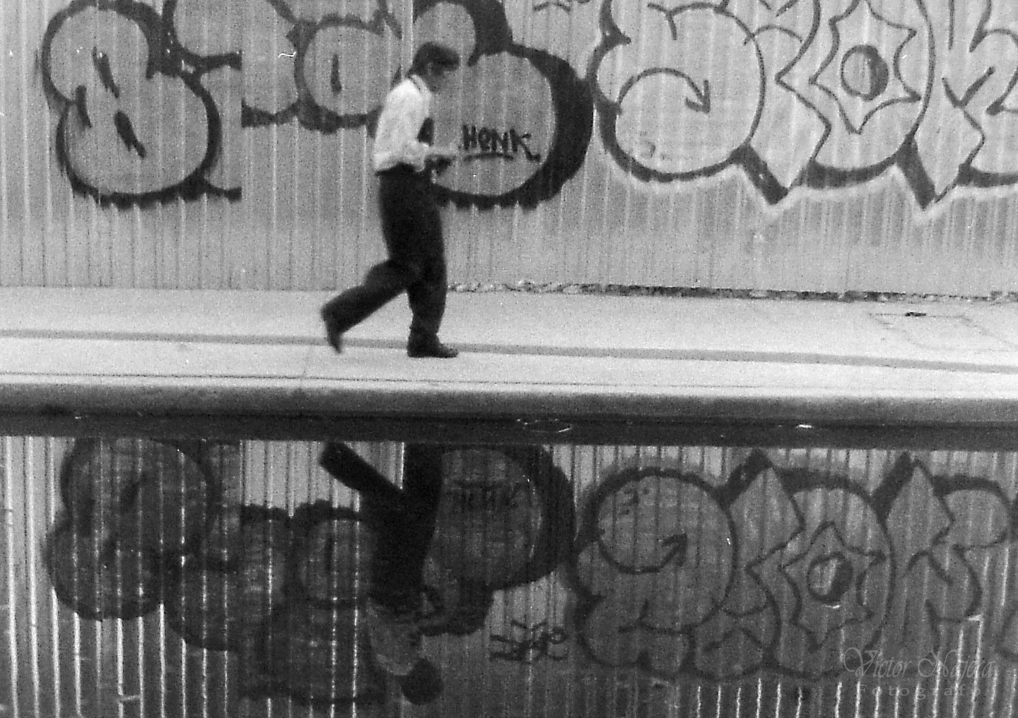A man walks along a sidewalk next to a graffiti-covered wall, with his reflection visible in a puddle below.