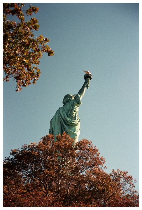 The Statue of Liberty is seen from behind, holding her torch high, with autumn trees in the foreground.