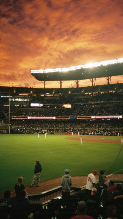 A baseball game is being played in a stadium under a dramatic orange and red sky.