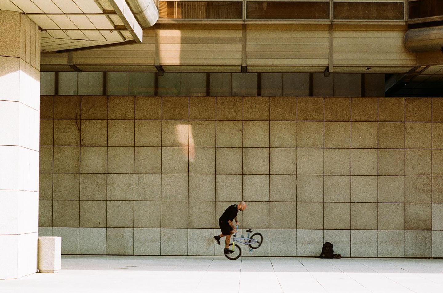 A man on a bicycle performs a trick against a tiled wall with sunlight and shadow.