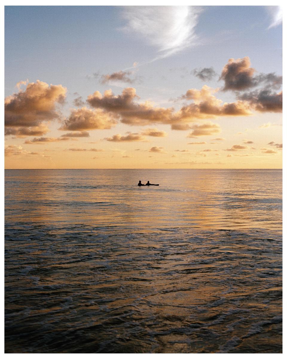 Two surfers are silhouetted against the ocean at sunset.