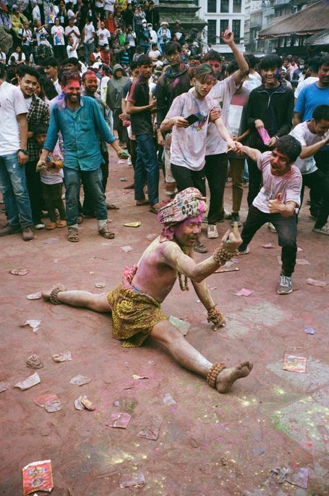 A man in a leopard print skirt and turban sits on the ground in a split with his arms raised, covered in colorful powder, while other people stand around him.