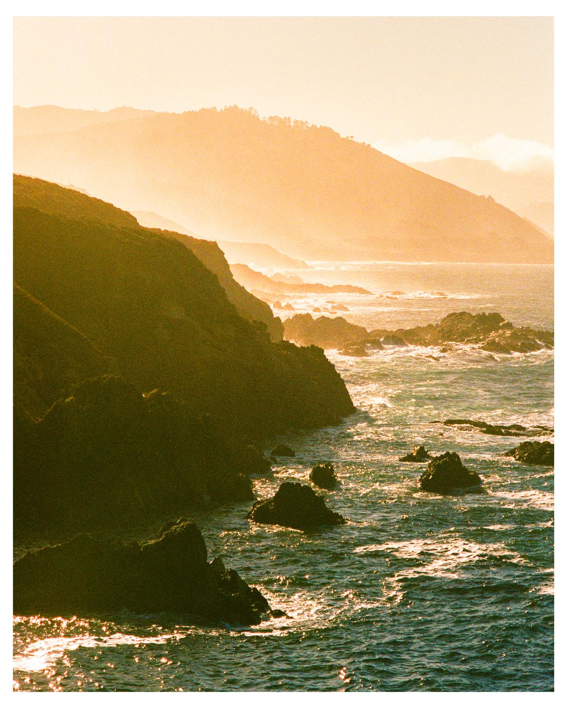 The sun shines on a rocky coastline with mountains in the background.