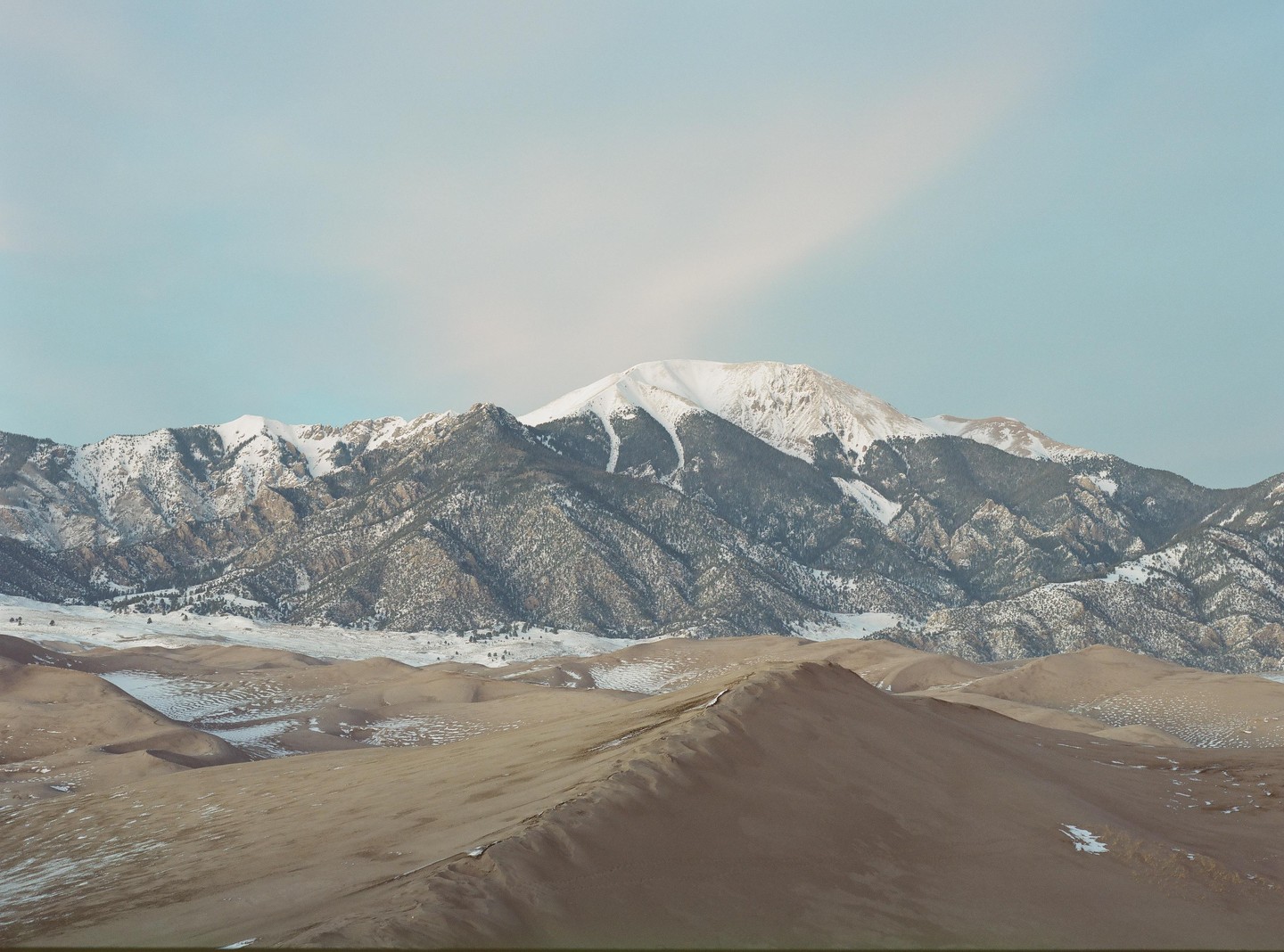A vast landscape shows sand dunes in the foreground and snow-capped mountains under a pale sky.