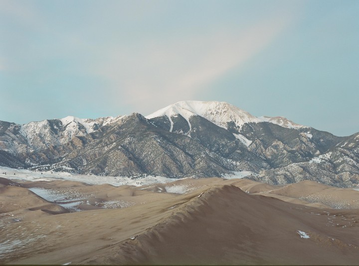 A vast landscape shows sand dunes in the foreground and snow-capped mountains under a pale sky.