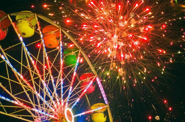 A ferris wheel with colorful gondolas is illuminated by fireworks exploding in the night sky.