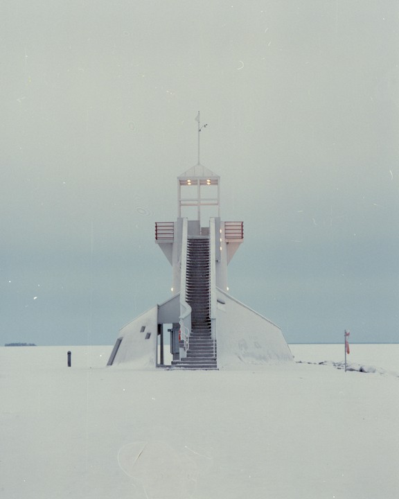 A white observation tower with a long staircase stands in a vast, snow-covered landscape under a pale sky.