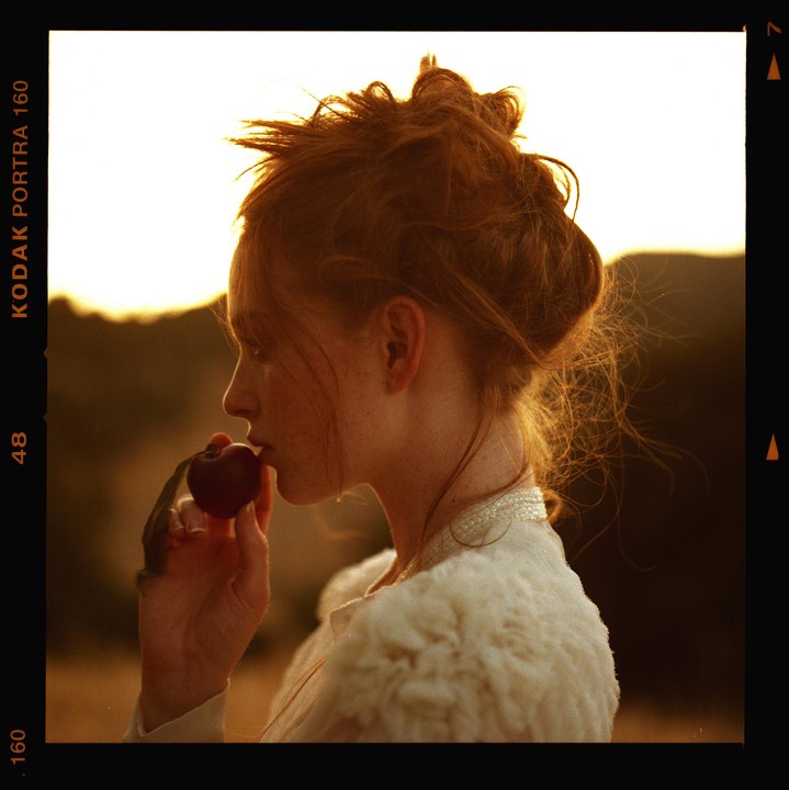A woman with red hair and freckles holds a plum to her lips.