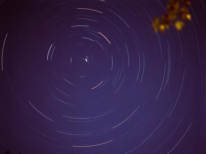 The night sky is filled with circular star trails around a central point, with some foliage visible in the upper right corner.