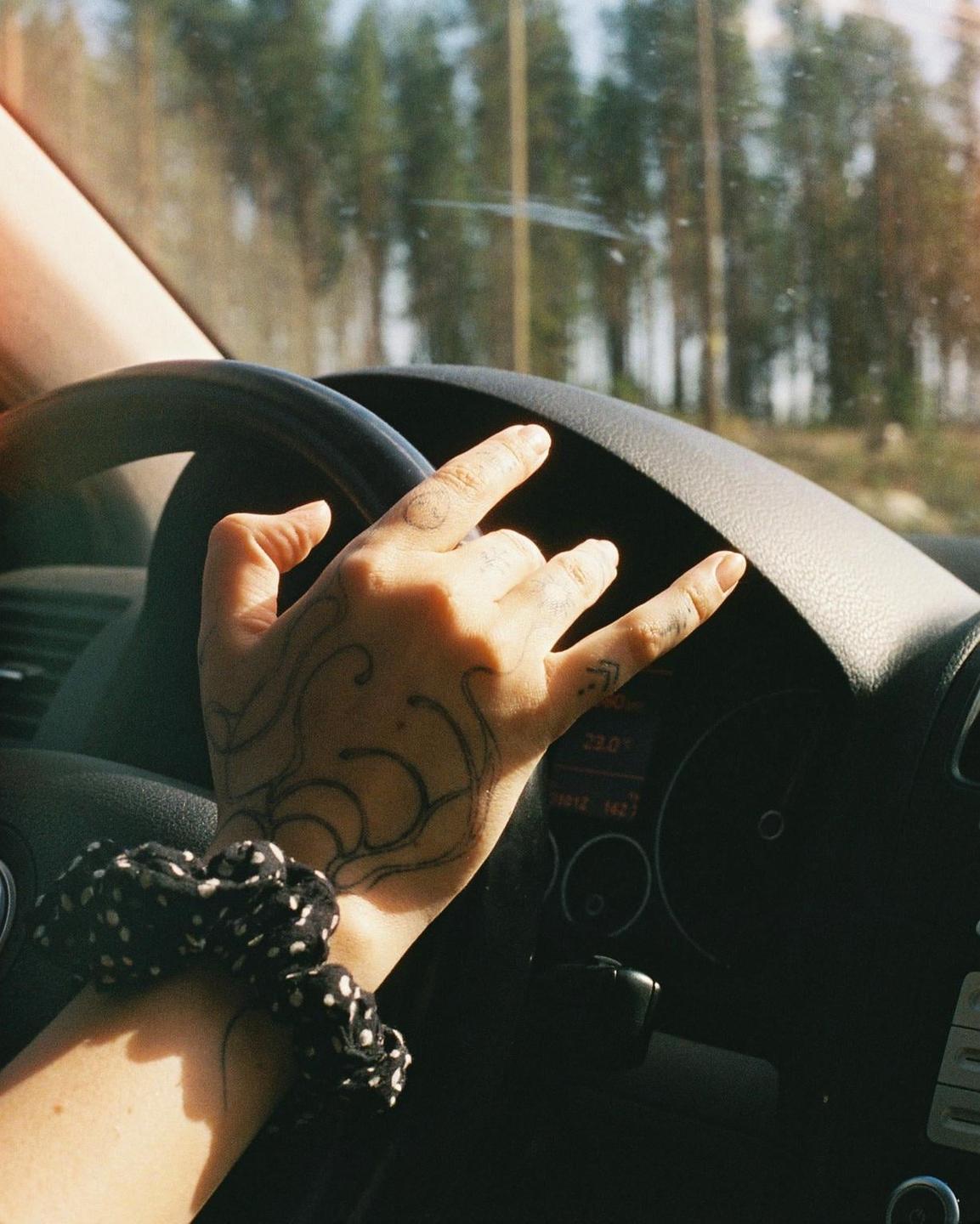 A tattooed hand with a scrunchie on the wrist makes a rock and roll gesture while driving a car.