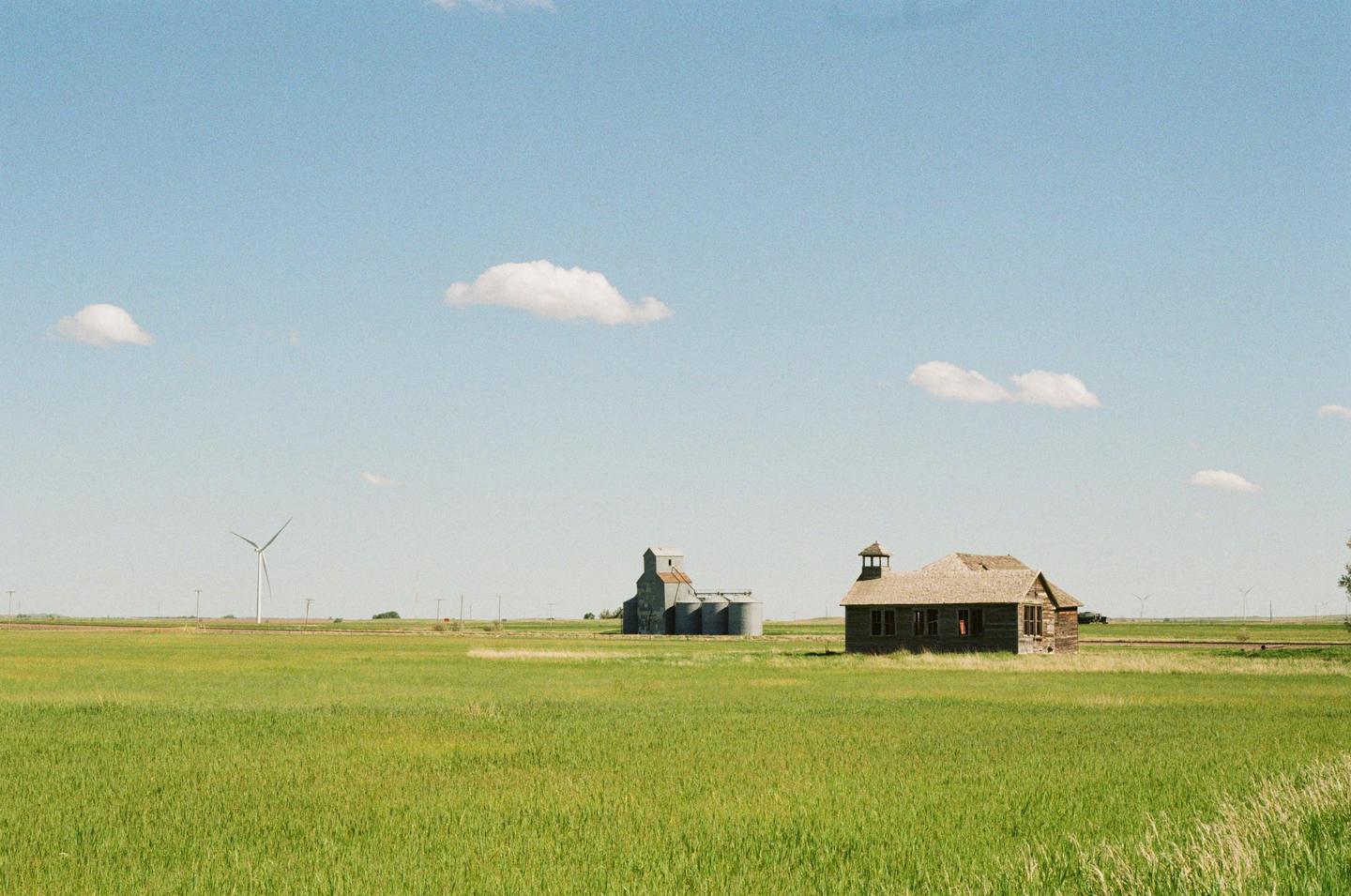 An old wooden building and a grain elevator stand in a green field under a blue sky with scattered clouds.