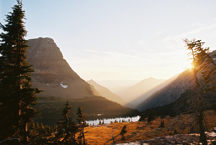 The sun sets over a mountain lake surrounded by pine trees and golden grass.