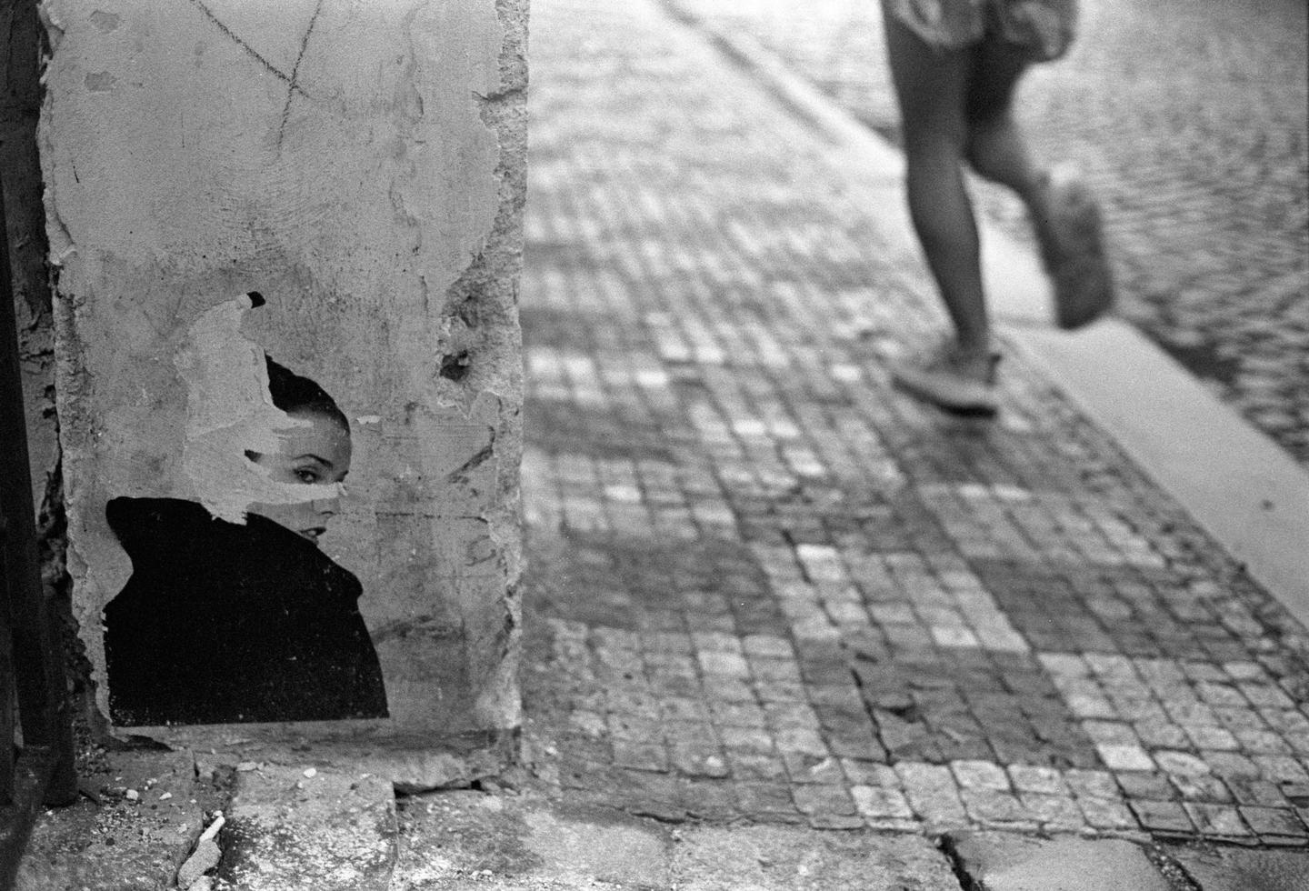 A torn poster of a woman's face is visible on a concrete wall next to a cobblestone street where a person is walking.