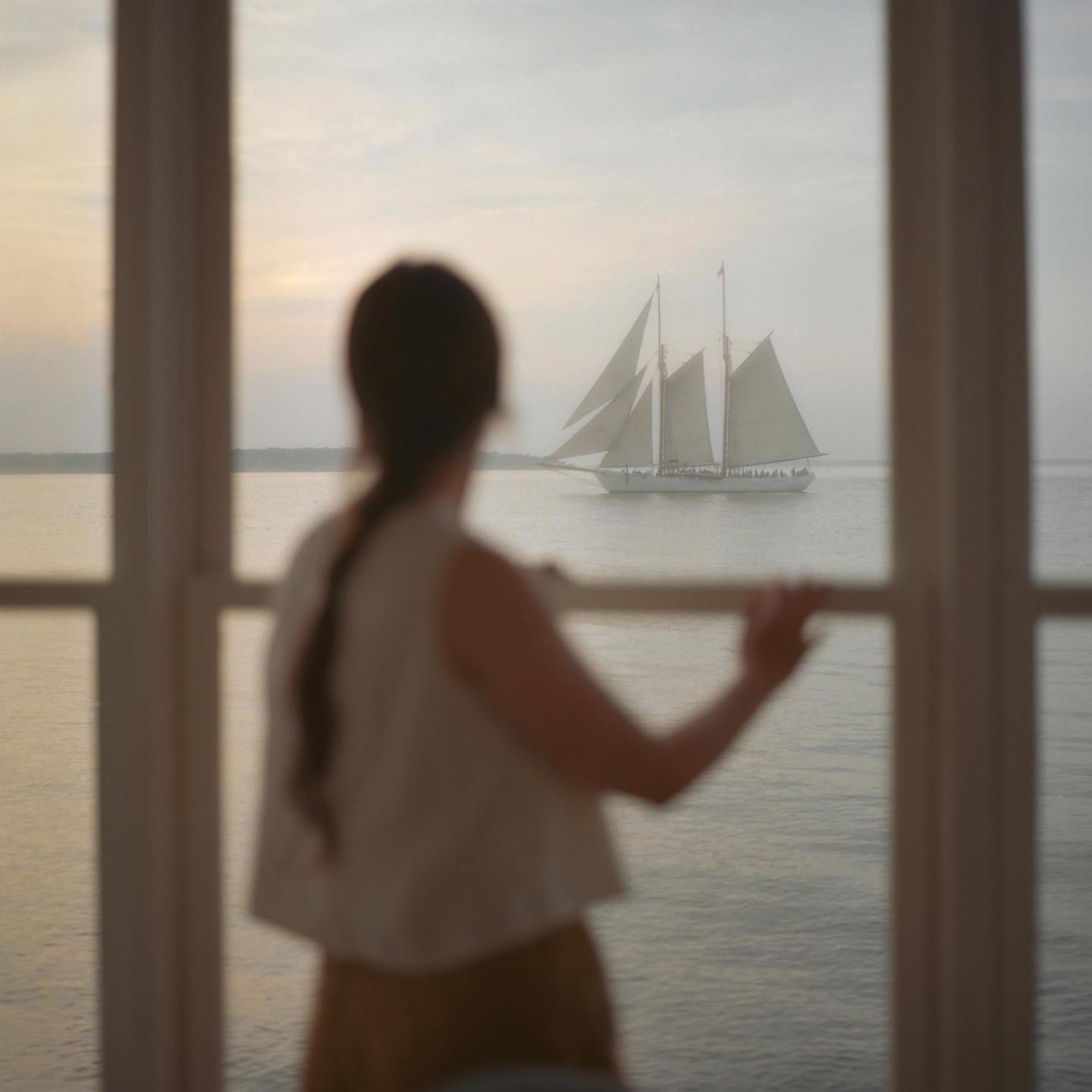 A woman looks out a window at a sailboat on the water at sunset.