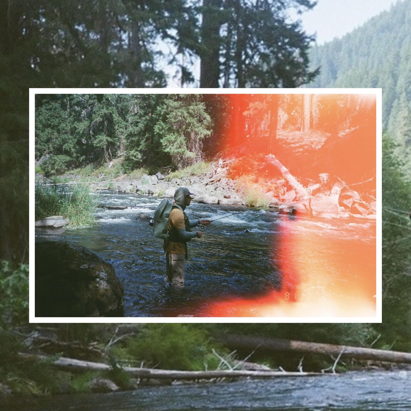 A man stands in a river, fishing with a rod, surrounded by trees and rocks.