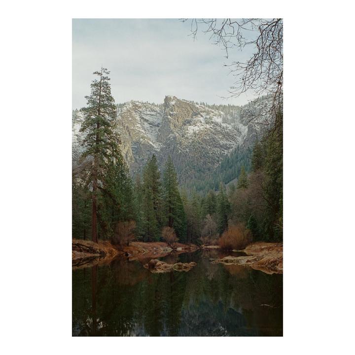 A river reflects a forest and snow-capped mountains under a cloudy sky.