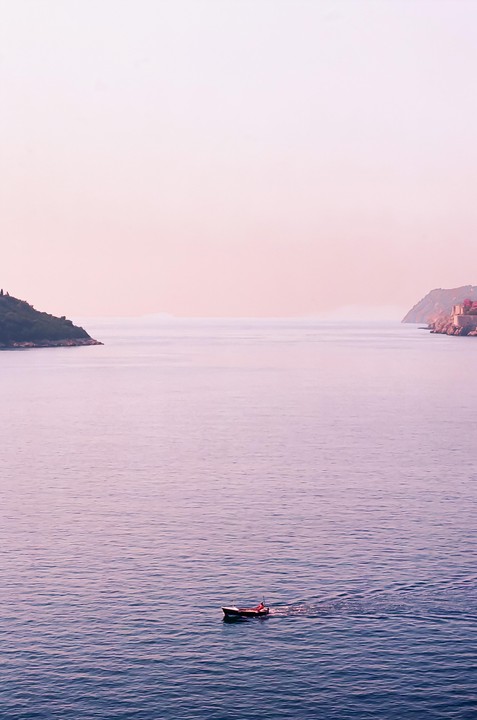 A small boat moves across calm water between two distant landmasses under a pink sky.