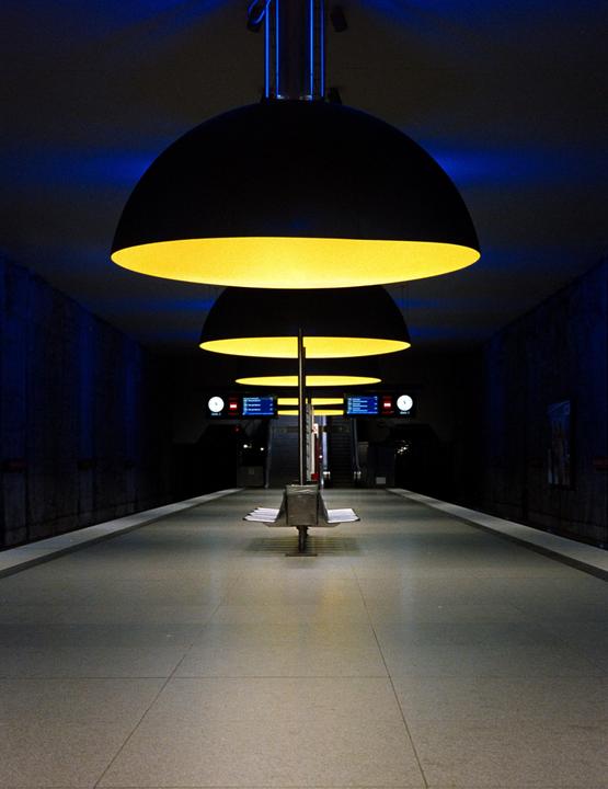 A subway platform is illuminated by a row of large, yellow, dome-shaped lights and blue ambient light.