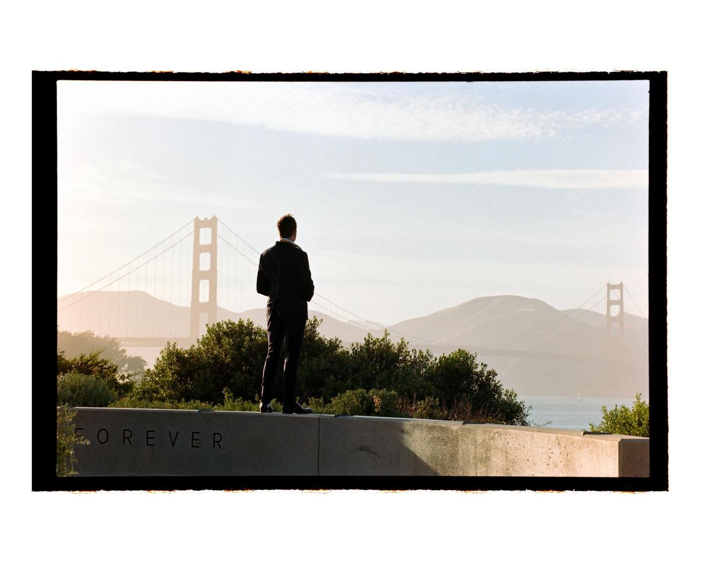 A man stands on a concrete wall looking out at the Golden Gate Bridge and a body of water with mountains in the distance.