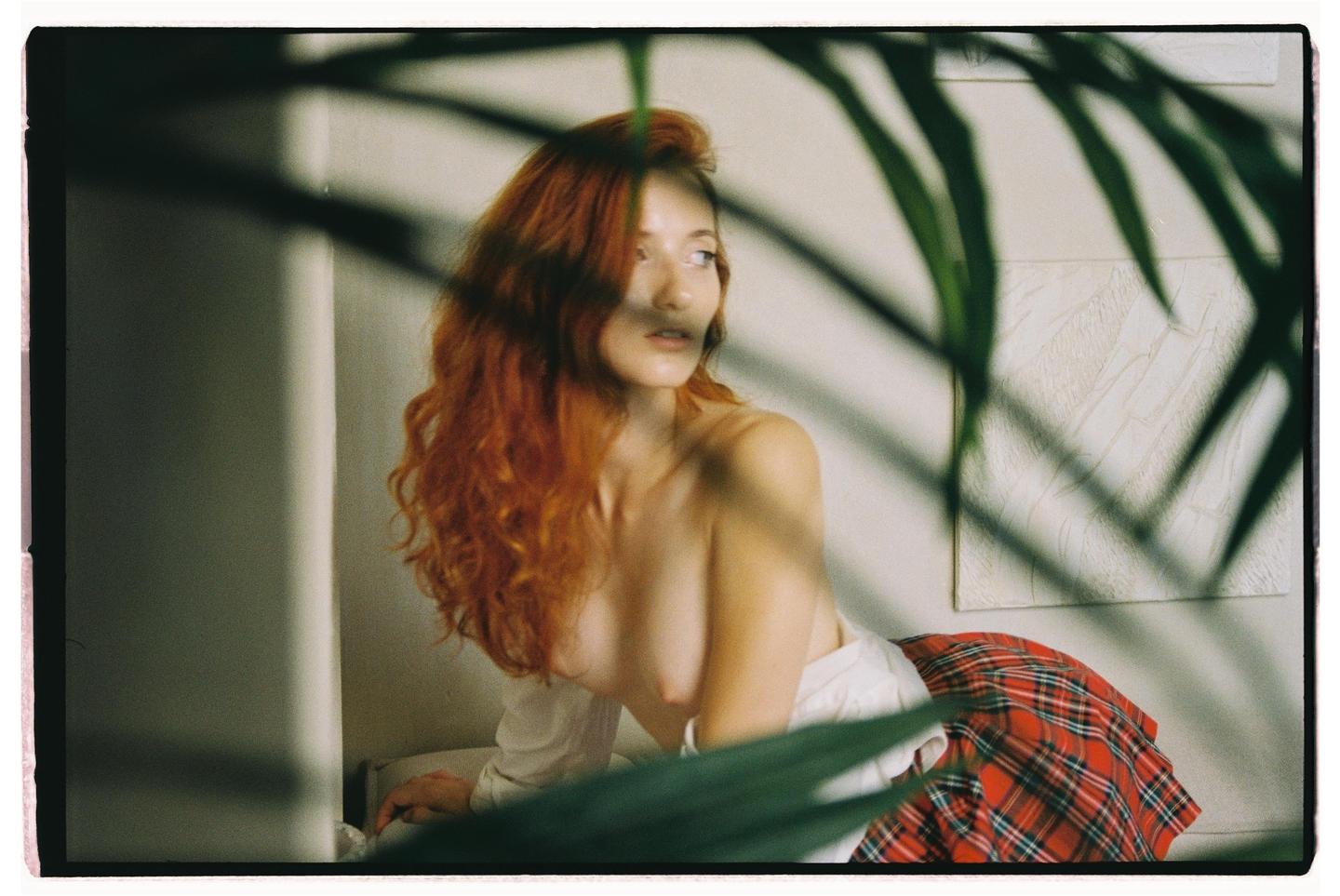 A woman with long red hair and a plaid skirt is partially obscured by plant leaves in the foreground.