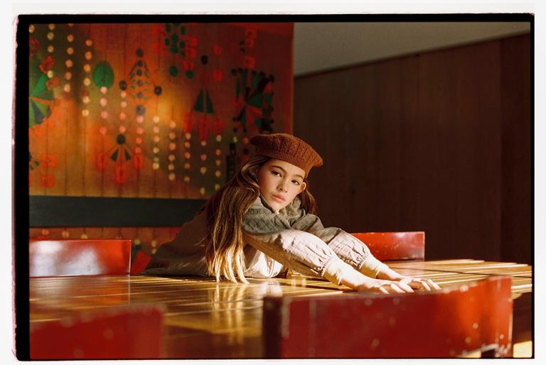 A girl in a brown beret lies on a wooden table with red chairs around her, illuminated by a warm light.