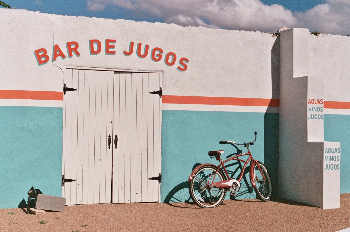 A red bicycle is parked in front of a white building with a blue and orange stripe and the words "BAR DE JUGOS" written on it.