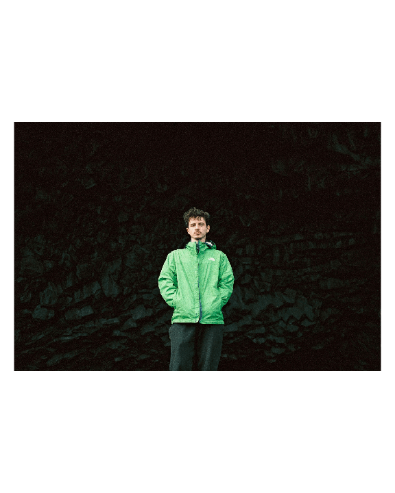 A man in a green jacket stands against a dark, rocky background.