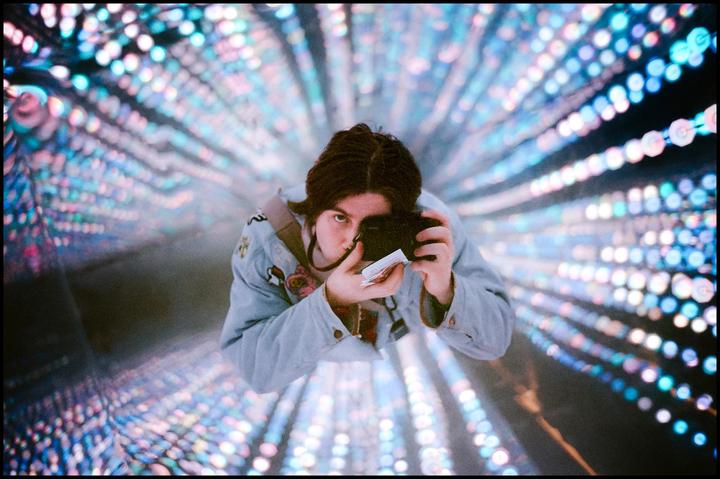 A woman holds a camera and looks up at the viewer, surrounded by a tunnel of colorful lights.