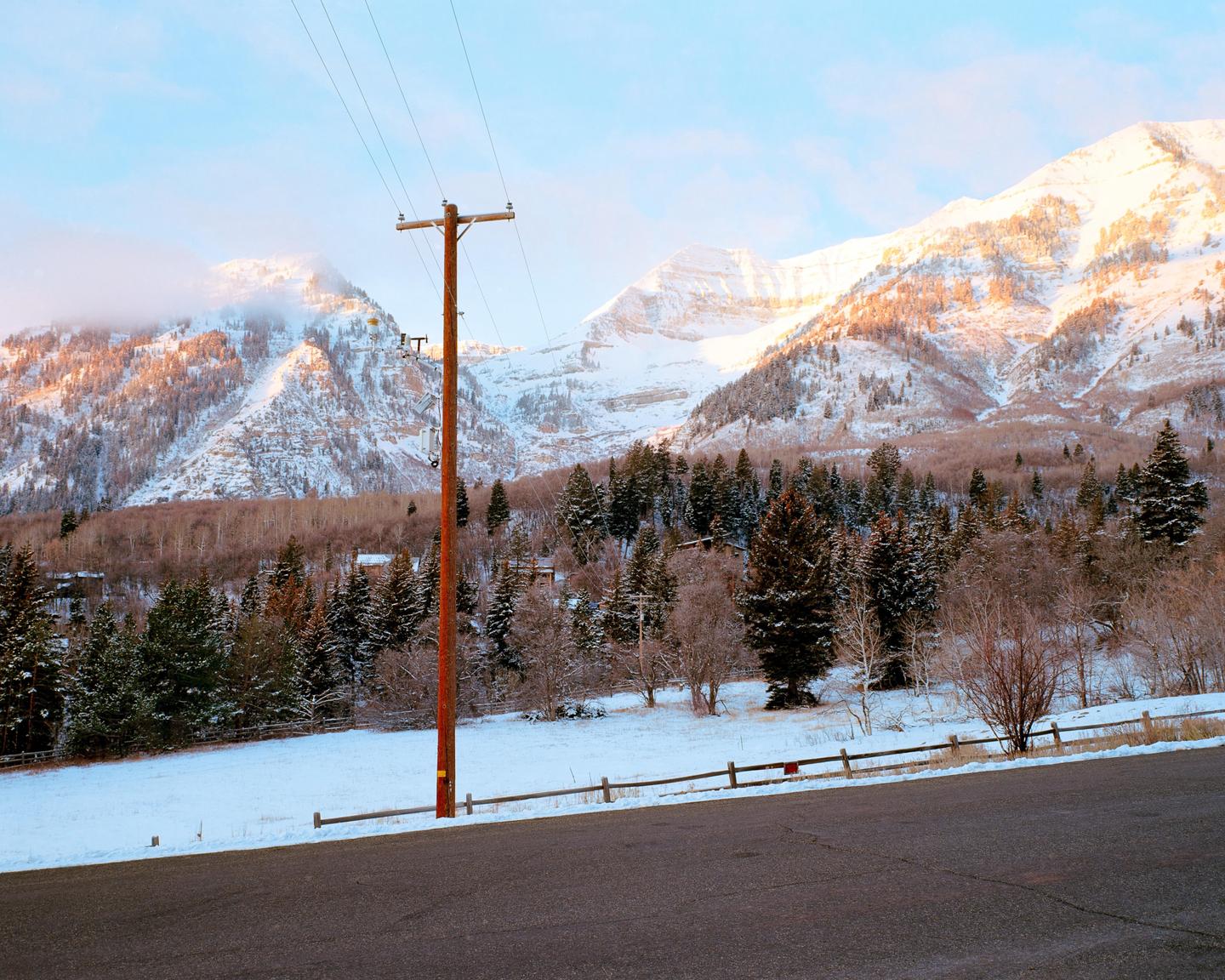 A snow-covered mountain range with pine trees and a utility pole in the foreground is illuminated by the setting sun.